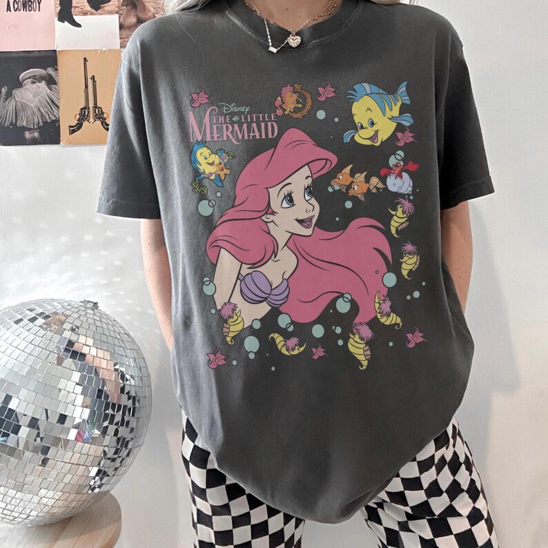 May include: Dark gray t-shirt featuring Ariel from Disney's The Little Mermaid. The graphic includes Ariel with pink hair, a purple seashell bra, and various sea creatures. The shirt has the movie title printed above the character.