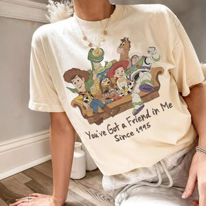 May include: A cream-colored short-sleeve t-shirt featuring a graphic print of Toy Story characters sitting on a couch. The text below reads You've Got a Friend in Me Since 1995. The design includes Woody, Buzz Lightyear, Jessie, Rex, Bullseye, and Slinky Dog.