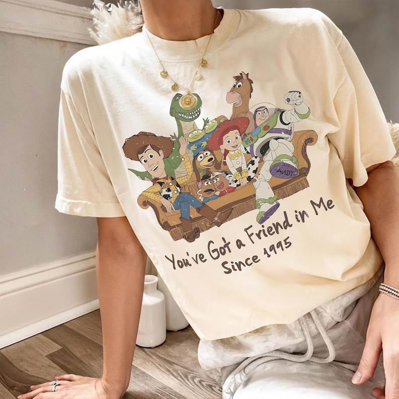 May include: A cream-colored short-sleeve t-shirt featuring a graphic print of Toy Story characters sitting on a couch. The text below reads You've Got a Friend in Me Since 1995. The design includes Woody, Buzz Lightyear, Jessie, Rex, Bullseye, and Slinky Dog.