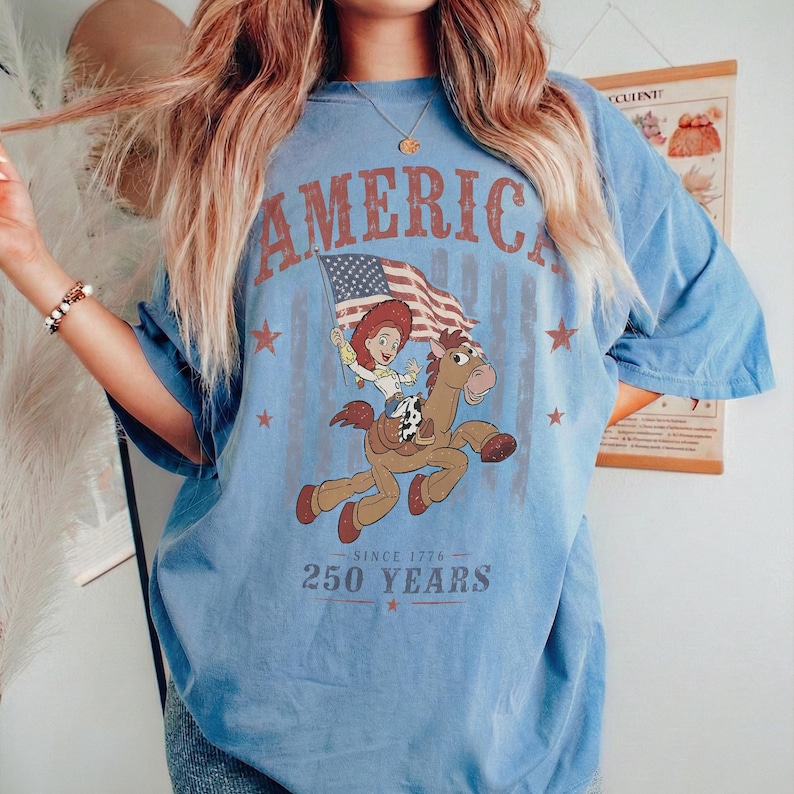 May include: A light blue t-shirt featuring a graphic of the character Jessie from Toy Story riding a horse while holding an American flag. The text reads AMERICA, SINCE 1776, and 250 YEARS. The design includes red stars and a faded flag background.