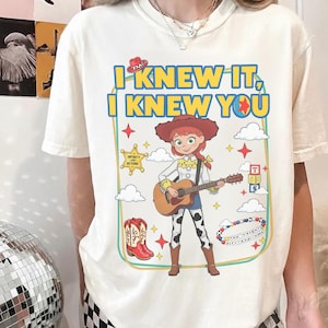 May include: A cream-colored short-sleeve t-shirt featuring a graphic of a cowgirl character playing an acoustic guitar. The text reads I KNEW IT, I KNEW YOU. The design includes a sheriff star, cowboy boots, alphabet blocks, and clouds.