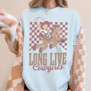 K&ouml;nnte beinhalten: Hellblaues Sweatshirt mit einem Cartoon-Cowgirl auf einem Pferd, mit Lasso, vor einem rot-wei&szlig; karierten Hintergrund. Der Text "LONG LIVE Cowgirls" ist unter der Grafik aufgedruckt.