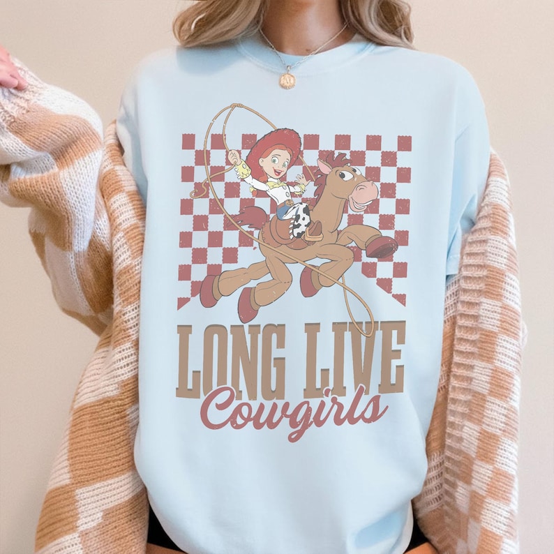 K&ouml;nnte beinhalten: Hellblaues Sweatshirt mit einem Cartoon-Cowgirl auf einem Pferd, mit Lasso, vor einem rot-wei&szlig; karierten Hintergrund. Der Text "LONG LIVE Cowgirls" ist unter der Grafik aufgedruckt.