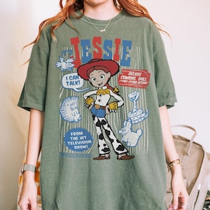 May include: An olive green t-shirt featuring a graphic of the character Jessie from the animated film Toy Story. The design includes the name "Jessie" in red and blue, along with the phrases "I Can Talk!" and "Deluxe Cowgirl Doll".