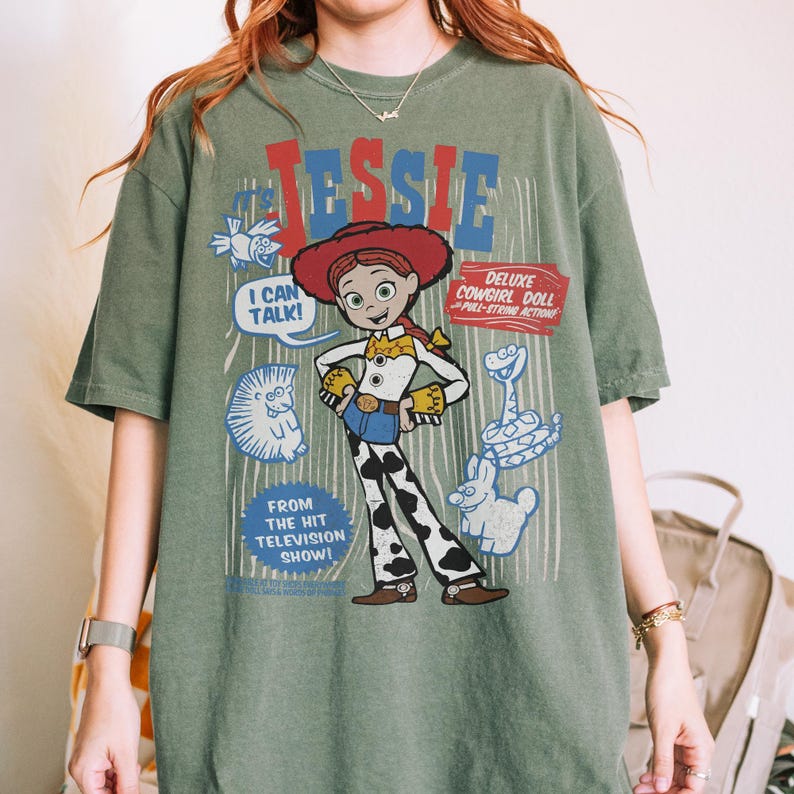 May include: An olive green t-shirt featuring a graphic of the character Jessie from the animated film Toy Story. The design includes the name "Jessie" in red and blue, along with the phrases "I Can Talk!" and "Deluxe Cowgirl Doll".