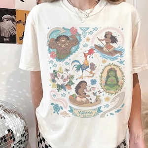 May include: Cream-colored t-shirt featuring colorful illustrations of characters from the animated film "Moana." The design includes images of Maui, Moana, Pua, Hei Hei, and other elements from the movie, with the text "Moana & Friends" at the bottom.