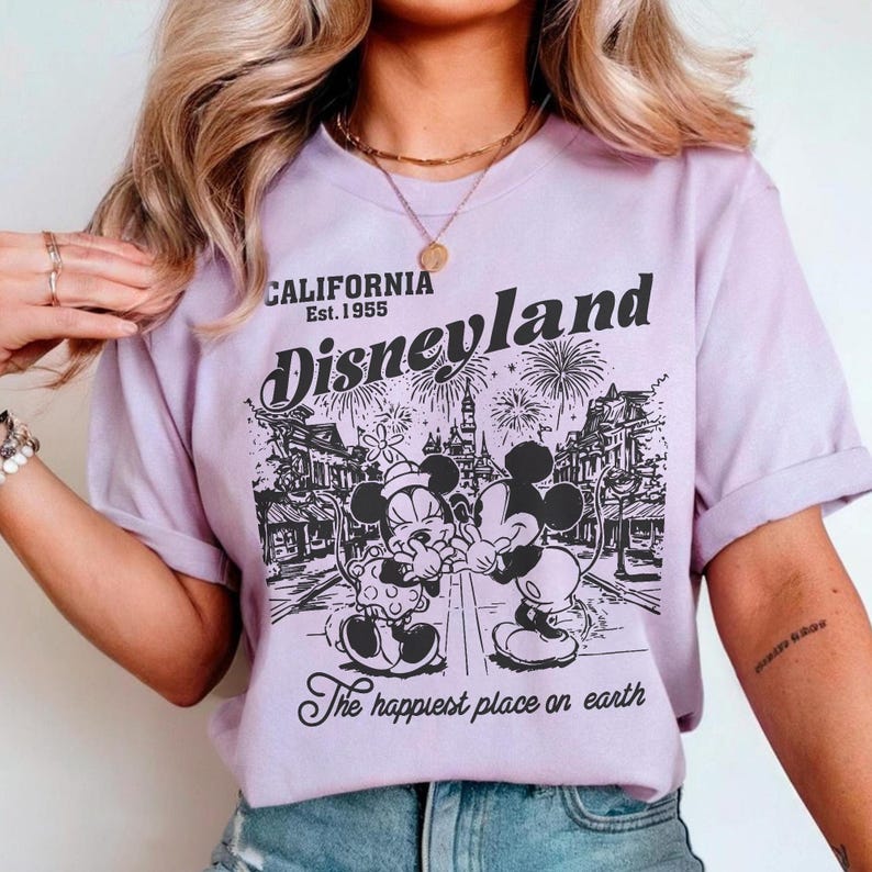 May include: A light purple t-shirt featuring a black and white graphic of Mickey and Minnie Mouse in front of a Disneyland scene with fireworks. The text "CALIFORNIA Est. 1955 Disneyland" and "The happiest place on earth" are also printed on the shirt.