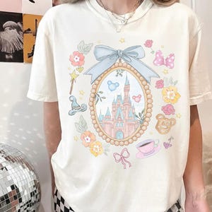 May include: Off-white t-shirt with a fairytale-themed design. A light blue bow adorns an oval frame showcasing a castle. The frame is surrounded by flowers, a magic wand, a witch's hat, a pink bow, a teacup, and a teddy bear.