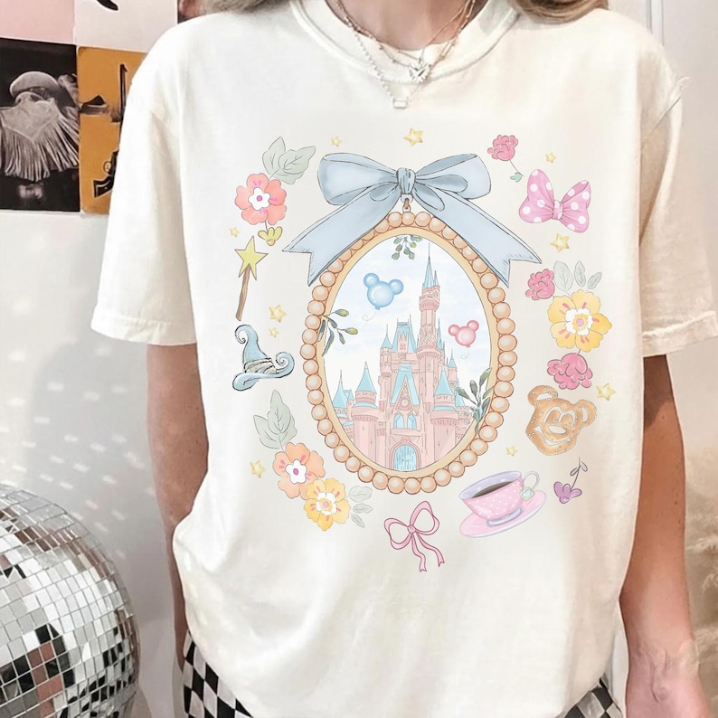 May include: Off-white t-shirt with a fairytale-themed design. A light blue bow adorns an oval frame showcasing a castle. The frame is surrounded by flowers, a magic wand, a witch's hat, a pink bow, a teacup, and a teddy bear.