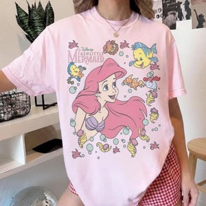May include: A light pink t-shirt featuring a graphic of Ariel from Disney's The Little Mermaid. Ariel has long pink hair and a purple seashell bra. The shirt also includes illustrations of Flounder, Sebastian, and other sea creatures.