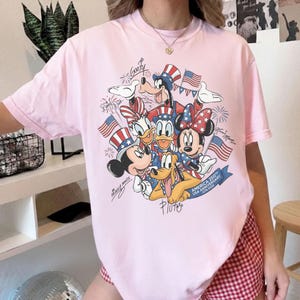 May include: A pale pink t-shirt with a graphic of Disney characters, including Mickey Mouse, Minnie Mouse, Donald Duck, Daisy Duck, Goofy, and Pluto, celebrating the USA's 250th anniversary. The design incorporates American flags and patriotic colours.
