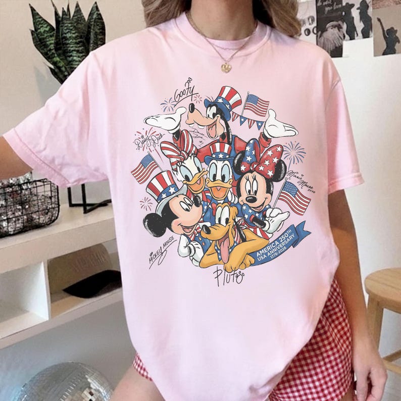 May include: A pale pink t-shirt with a graphic of Disney characters, including Mickey Mouse, Minnie Mouse, Donald Duck, Daisy Duck, Goofy, and Pluto, celebrating the USA's 250th anniversary. The design incorporates American flags and patriotic colours.
