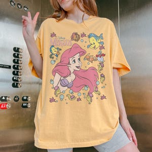 May include: Yellow t-shirt featuring a graphic of Ariel from Disney's The Little Mermaid. The design includes Ariel with pink hair, a purple seashell bra, and various sea creatures. The text "Disney The Little Mermaid" is also visible.