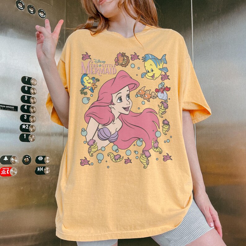 May include: Yellow t-shirt featuring a graphic of Ariel from Disney's The Little Mermaid. The design includes Ariel with pink hair, a purple seashell bra, and various sea creatures. The text "Disney The Little Mermaid" is also visible.