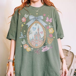May include: Olive green t-shirt with a framed castle illustration, surrounded by flowers, bows, and a teacup. The design includes a light blue bow and a pink bow. The shirt has a loose fit and is made of a soft material.