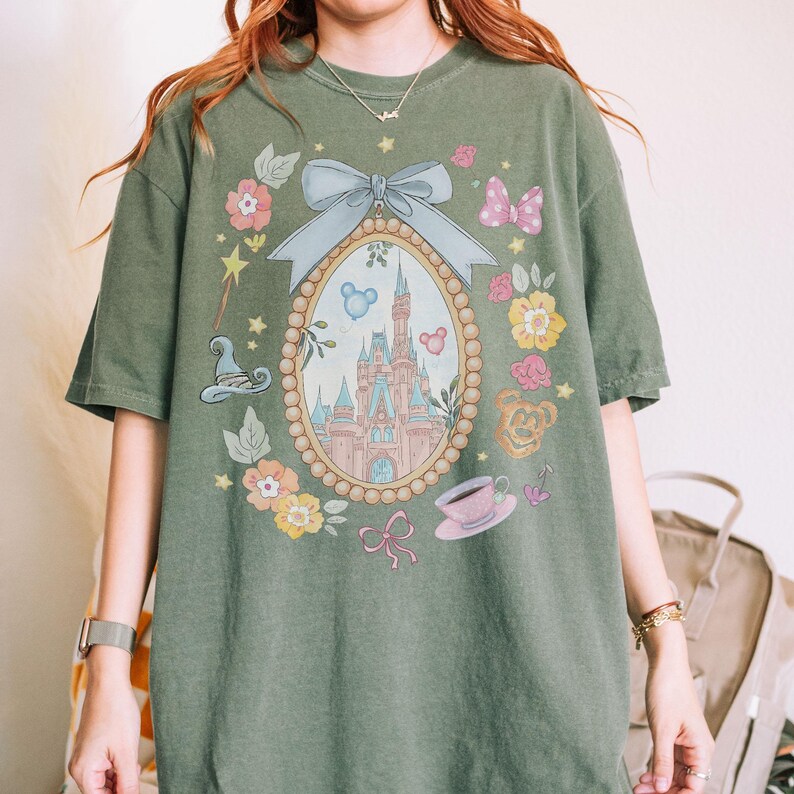 May include: Olive green t-shirt with a framed castle illustration, surrounded by flowers, bows, and a teacup. The design includes a light blue bow and a pink bow. The shirt has a loose fit and is made of a soft material.