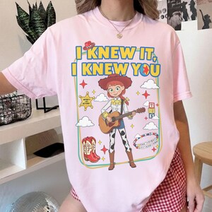 May include: A light pink t-shirt featuring a graphic print of a cowgirl character playing an acoustic guitar. The text reads I KNEW IT, I KNEW YOU. The design includes a cowboy hat, a star badge, cowboy boots, alphabet blocks, and clouds.