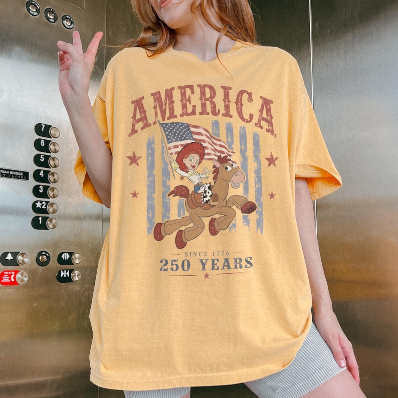 May include: A yellow short-sleeve t-shirt featuring a graphic of a cowgirl riding a horse with an American flag. The text reads AMERICA at the top and SINCE 1776 250 YEARS at the bottom, accented by red stars and faded blue vertical stripes.