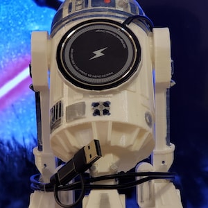 Puede incluir: Un cargador inalámbrico R2-D2 blanco y azul con una base de carga negra. La base de carga tiene un símbolo de rayo y texto. Un cable USB negro está conectado al cargador.