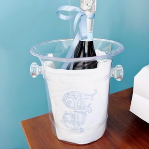 Puede incluir: Una cubeta de champán de plástico transparente con dos asas contiene una botella de champán. La botella está envuelta con una cinta azul claro. La cubeta está forrada con un paño blanco bordado con un monograma azul claro.