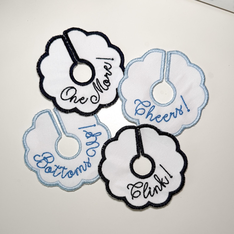 May include: Four white, scalloped-edge drink charms with embroidered text. The charms have a split design to wrap around a glass stem. Text includes "One More!", "Cheers!", "Bottoms Up!", and "Clink!" in black and blue thread.