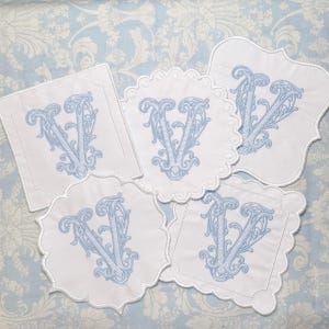 May include: Five white coasters with a light blue embroidered letter "V" in a decorative script. Each coaster has a different scalloped or shaped edge. The coasters are arranged on a light blue floral patterned fabric.