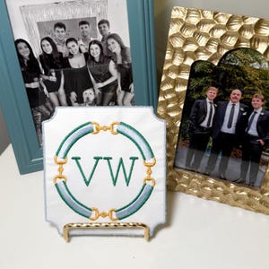 May include: White embroidered square with green and gold accents, featuring the letters "VW". The design includes a circular pattern with a horse bit motif. The square is displayed on a gold stand, with framed photos in the background.