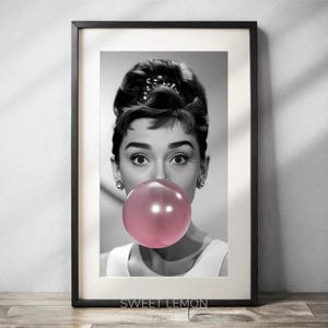 May include: Black and white photo of a woman with a large pink bubble gum in her mouth. The woman is wearing a white shirt and has her hair styled in a bun. The photo is framed in a black frame.