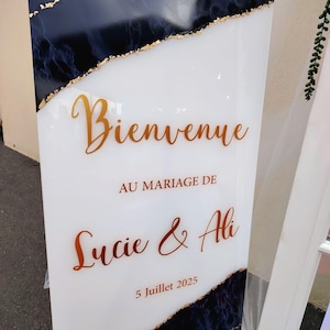 Peut inclure: Panneau de bienvenue de mariage blanc avec lettrage et accents dorés. Il est écrit "Bienvenue Au Mariage De Lucie & Ali 5 Juillet 2025". Bordures supérieures et inférieures en marbre bleu foncé et feuilles d'or.