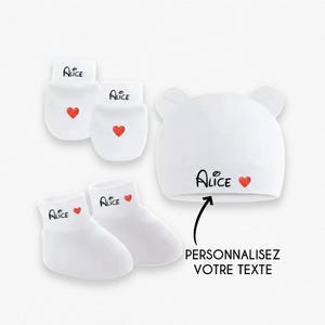 Può includere: Set bianco per neonati con guanti, calzini e cappello con il nome "Alice" e un cuore rosso. Il cappello ha le orecchie da orso. È presente anche il testo "PERSONNALISEZ VOTRE TEXTE".