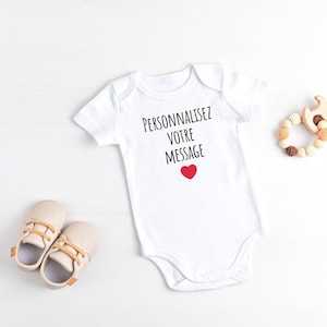 Peut inclure: Un body blanc pour bébé avec le texte "Personnalisez Votre Message" et un cœur rouge. Le body est posé à plat sur une surface blanche avec un jouet en bois et des chaussures de bébé.