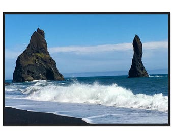 May include: A landscape photograph of a black sand beach with two large, dark, jagged rock formations rising from the blue ocean. White sea foam crashes against the shore under a clear blue sky with a thin horizontal cloud formation.