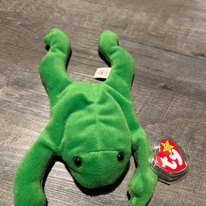 May include: Green frog beanie baby with a red and white Ty tag. The tag has a star and the words "Beanie Babies" on it.