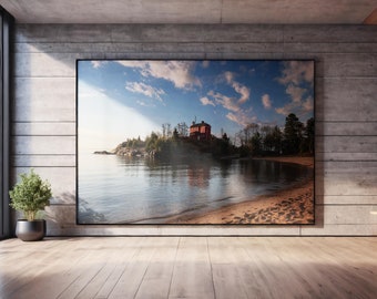 May include: A large rectangular wall art print featuring a landscape photograph of a red building situated on a rocky shoreline by a calm body of water under a blue sky with scattered clouds, displayed in a thin black frame on a concrete wall.