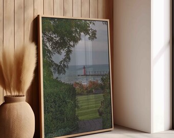 May include: A framed print of a coastal scene featuring a red lighthouse at the end of a pier. The artwork is set against a backdrop of a cloudy sky and water, framed by lush greenery and a manicured lawn. The frame is a light wood color.