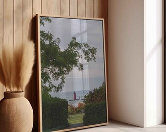 May include: A framed landscape print featuring a red lighthouse on the coast, framed by green foliage and a cloudy sky. The print is in a light wood frame, leaning against a wooden wall. A beige vase with dried pampas grass is in the foreground.
