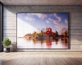 May include: A large rectangular wall art print featuring a vibrant landscape photograph of a red lighthouse situated on a rocky island surrounded by water, with a bright blue sky and scattered clouds, displayed in a black frame on a concrete wall.