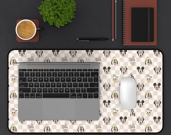 May include: A desk mat featuring a repeating pattern of cartoon character faces on a checkered background. A laptop, mouse, and other office supplies are on the mat. A cup of coffee and a small plant are also on the desk.