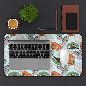 May include: A light blue mousepad with a pattern of cartoon castles, trees, and geometric shapes. A laptop, white mouse, and other office supplies are on the black surface. A cup of coffee and a small plant are also visible.