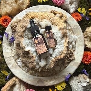 May include: Two black glass bottles of "Star Magic" oil with labels featuring an owl and star design. The bottles are on a stone surface with crystals, flowers, and moss. One bottle has a dropper, the other a small cap.