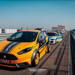 May include: A group of race cars on a track. The leading car is a vibrant orange with black stripes and accents, displaying "FONDA" on its number plate. Other cars follow, with a clear blue sky above.