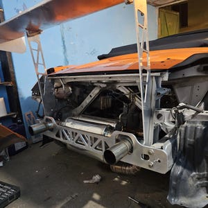 Pode incluir: Um carro desportivo laranja em modificação, com uma grande asa traseira prateada e um sistema de escape personalizado. A carroçaria do carro está parcialmente desmontada, revelando o compartimento do motor e componentes estruturais. Numa garagem.