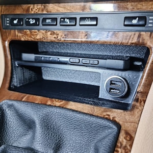 May include: A close-up of a car's interior with a wooden dashboard and a black phone holder with two USB ports. The phone holder is located in the center console, below the buttons for the car's features.