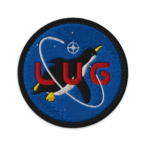 May include: Embroidered patch featuring a black and white penguin with yellow feet and beak, flying through a white arc on a blue background. The red letters "LUG" are prominently displayed. The patch has a black border.