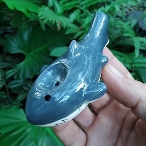 May include: A gray ceramic shark pipe with a white underbelly and black eye. The pipe has a round hole on the top for smoking.