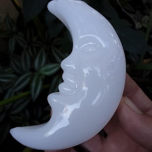 May include: A white, crescent moon-shaped sculpture with a serene face carved into its surface. The sculpture is smooth and glossy, reflecting light. The background features green foliage, enhancing the moon's bright appearance. The sculpture is held in a hand.
