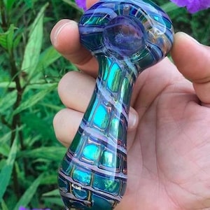 May include: A hand-blown glass pipe with a vibrant, iridescent design. The pipe features a teal, blue, and purple color scheme with a grid-like pattern. The bowl is round, and the stem is teardrop-shaped. The background includes green foliage and purple flowers.