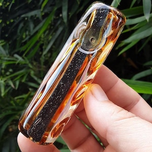 May include: A clear glass pipe with a rectangular shape. The pipe features a dark, speckled interior and is accented with swirls of amber, white, and gold. The design includes a circular opening on the side.