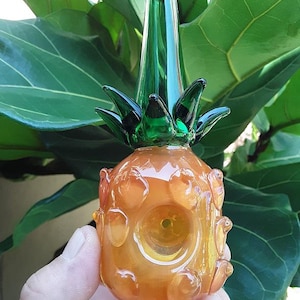 May include: A glass pineapple-shaped pipe with a green top and a clear bowl. The pipe is made of clear glass with a textured surface.