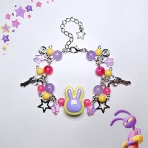 Puede incluir: Una colorida pulsera con un dije de conejito, dijes de estrella y mariposa, y dijes de pistola. La pulsera incluye cuentas rosas, amarillas y moradas, y está conectada por una cadena plateada. Sobre un fondo blanco.